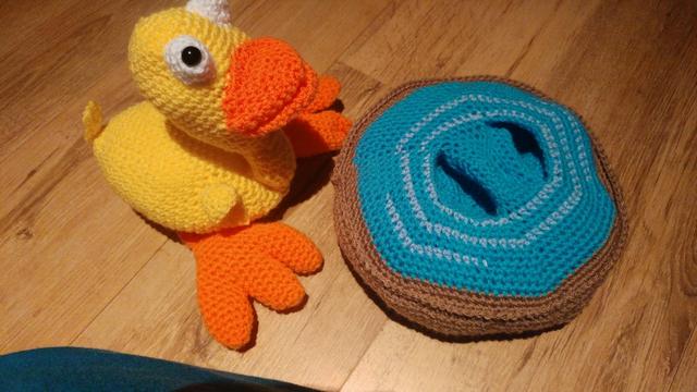 The same yellow crochet duck now out on the floor next to the crochet puddle. His large orange webbed feet are now visible and two hollows in the crochet puddle are now visible. This is where his feet are tucked into when he sits on the puddle.