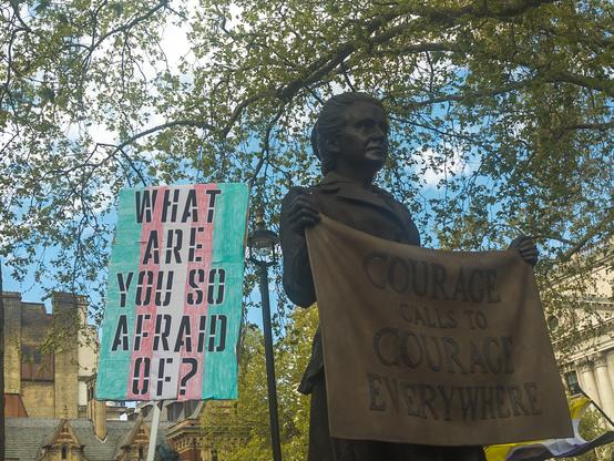 A protest placard reads "WHAT ARE YOU SO AFRAID OF" stencilled atop the colours of the transgender flag, it's next to a statue of a woman holding a fabric sign saying "Courage calls to courage everywhere"