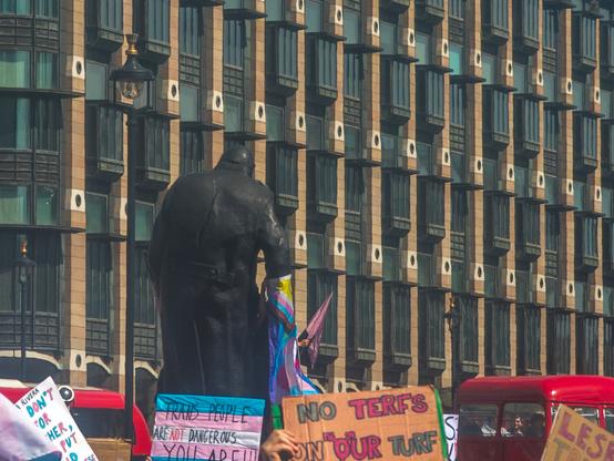 A statue of Winston Churchill has a progress pride flag around one arm. Below are protest signs including "Trans people are not dangerous YOU ARE" and "No TERFs on our turf"