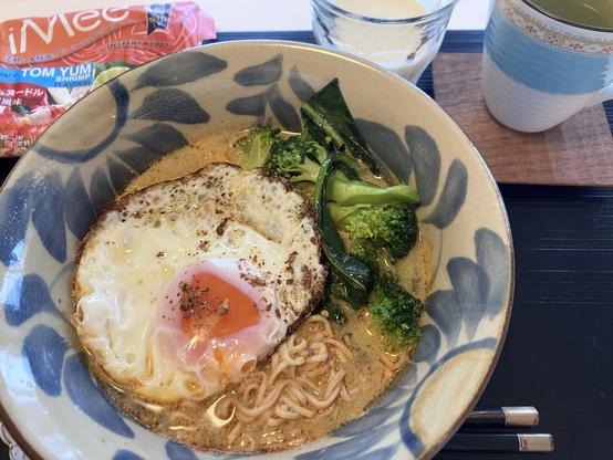 A bowl of creamy Tom Yum instant noodles topped with a fried egg, alongside broccoli and leafy greens. In the background, there is a glass of a dairy beverage and a cup of green tea on a wooden tray.