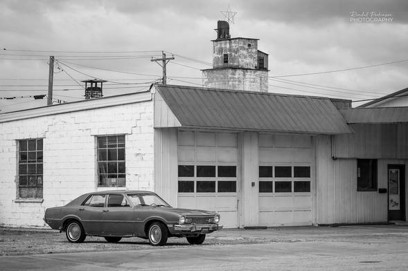 Black and white image of a 1970's Ford Maverick automobile sitting in front of an old auto repair buidling with two garage doors.