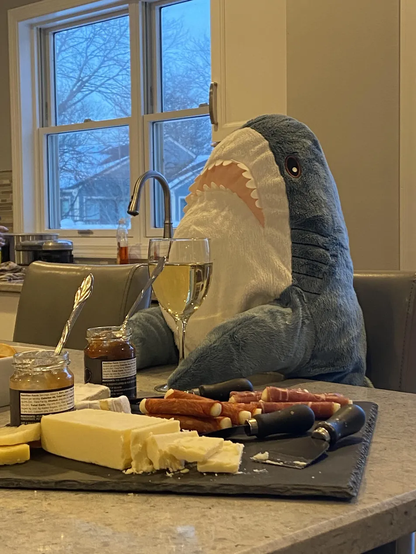 A BLÅHAJ shark plushie seated at a kitchen counter enjoying what appears to be a sophisticated charcuterie board. The blue and white shark is positioned upright beside a slate serving board loaded with cheese, prosciutto-wrapped breadsticks, and jars of what look like preserves or spreads with serving spoons. A glass of white wine sits next to the shark, completing the elegant snack setup. Through the windows in the background, a snowy winter scene is visible with bare trees, suggesting a cozy indoor gathering during cold weather. The scene humorously portrays the stuffed toy as if it's having a refined dining experience.