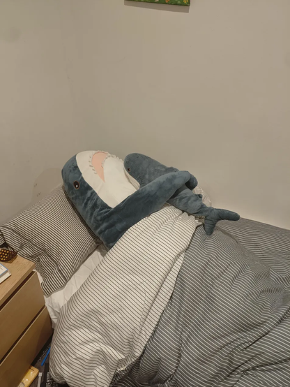 Two BLÅHAJ shark plushies in bed together, arranged to appear as if they're cuddling. One shark has its fin resting over the other, creating a sweet embracing pose. They're tucked in under gray and white striped bedding on what looks like a single bed with matching striped pillows. A wooden bedside table or drawer is visible on the left side. The sharks' distinctive blue bodies, white bellies, and pointed fins are clearly visible against the minimalist bedroom setting with white walls. The scene creates a cozy, endearing tableau of the popular IKEA sharks sharing a restful moment together.