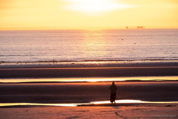 A beach at low tide against the backdrop of a setting sun. In the midground, a single person stands as a silhouette, surrounded by shimmering water from tidal pools that stretch towards the open sea. On the horizon, a distorted silhouette of a container ship is visible on the water, with certain areas appearing exaggerated in height.