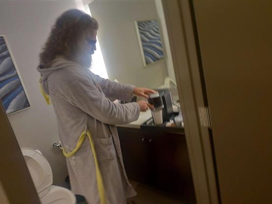 A large, hairy man, wearing a My Little Pony bathrobe, tennis shoes, and sunglasses, making coffee in a hotel bathroom.