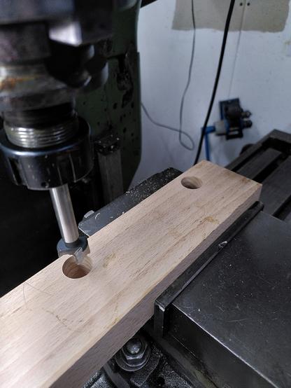 A block of beech in the vice of a metal machining mill.the beech has two holes drilled into the top face. A 20mm Forstner bit is visible on the left of the shot.
