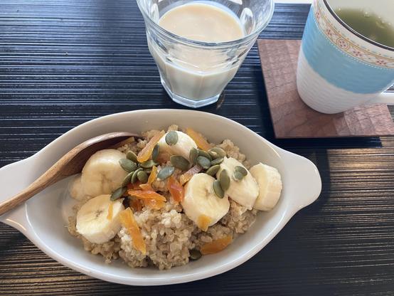 A bowl of quinoa topped with banana slices, pumpkin seeds, and dried apricots, accompanied by a glass of milk and a cup of green tea on a wooden coaster. The setting features a black surface.
