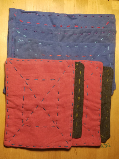 Photo of many finished zokin- rectangular cleaning clothes made from old tshirts in red, grey, & blue. There is blue, red, purple, or pink embroidery thread forming an X & a series of concentric boxes on each of them.