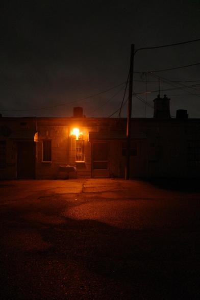 Color photograph looking at the rear entrance of a building at night. A security light shines an orange-ish light over an empty parking lot, somewhat cracked, and along the rear face of an old commercial building, a utility pole stretches up above the top of the building, silhouetted along with some power lines and chimneys against an overcast sky