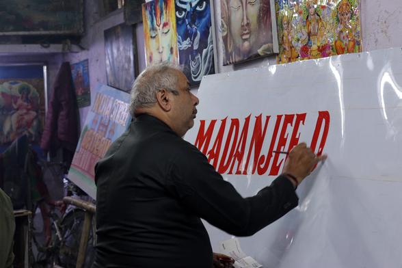 Man in a artistic studio setting painting red lettering onto a white surface.