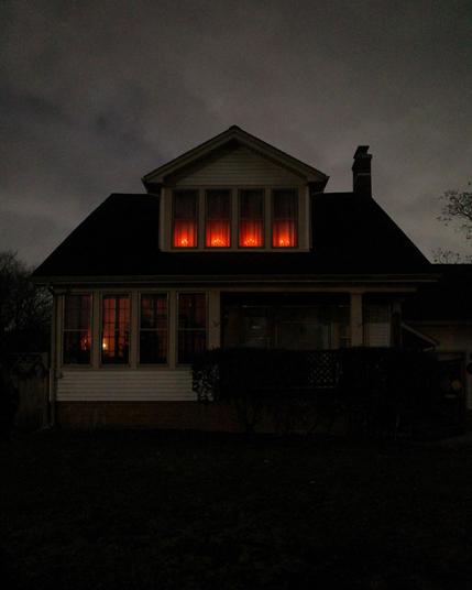 Color photograph looking at the darkened front of a house on an overcast night, orange lights from decorative pretend candles shine in four narrow windows across a large second-floor gable, and the rest of the house is in deep shadow or silhouetted against the bright slate gray sky