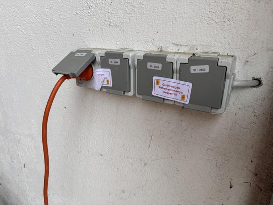 Ein grauer Mehrfachsteckdosenblock an einer Wand. Eine orange Verlängerungkabel ist an eine der Steckdosen angeschlossen. Auf einer Steckdose befindet sich ein Aufkleber mit der Aufschrift "Gerät wegen Sicherheitsmängel gesperrt".