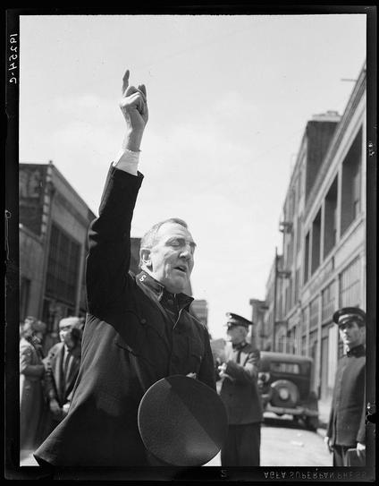 The black and white photograph captures a moment of public exhortation or speech, likely taken during the 1930s given its style. A man in military-style attire is seen raising his right hand towards the sky with fingers extended upwards as if making an emphatic point. He appears to be addressing a crowd out-of-frame; several individuals can be observed behind him, including what seems like a police officer on the far left and other men possibly listening or participating in the event.

The setting is urban with industrial buildings lining either side of a street where two vintage cars are parked—one facing towards us and another further down the road. The sky above is clear but has no discernible features to suggest weather conditions, time of day, or specific geographic location beyond that it's daytime outdoors within an American city.

The man's expression exudes confidence and conviction, suggesting he may be a figure of authority such as a public speaker, politician, or community leader. His attire indicates formality yet practicality for what might have been considered outdoor work during the era—indicative perhaps of wartime efforts given historical context clues like his cap bearing insignia.

The presence of military-style garments suggests this could relate to some aspect of World War II morale boosting activities as common in many Western countries at that time. The Salvation Army's involvement adds another la [...]