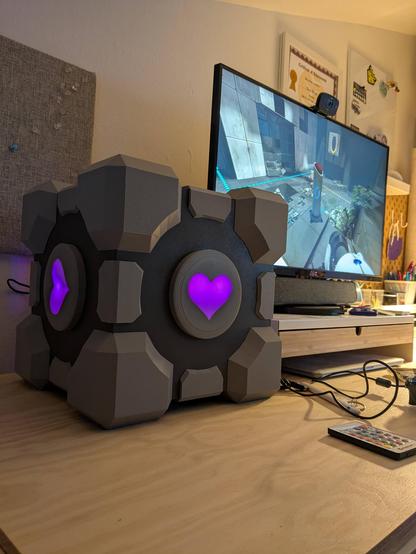 In the foreground, a computer case in the shape of a cube with projecting corners and edges and central heart cutouts which glow purple/pink. Behind, a computer monitor with a scene from the video game Portal.
