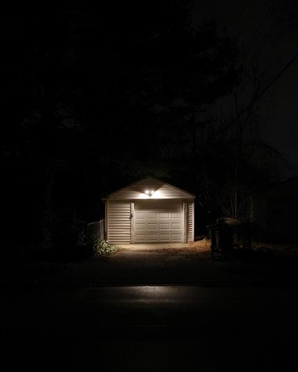 Color photograph of a standalone single-car garage on a dark night with a bright security light shining onto the face of the garage and driveway from above the closed garage door, the rest of the fame is very dark, you can kind of make out parts of a fence and trees and stuff