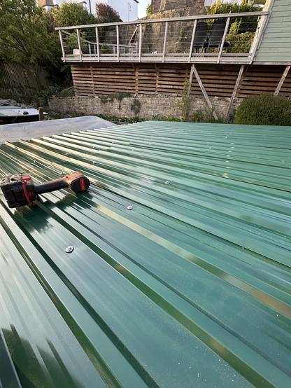 The green metal roof sheets of my woodstore seen from above. An impact driver is lying on its side.