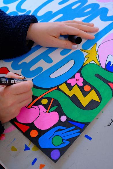 A person uses markers to color a vibrant, abstract drawing featuring bold shapes, hands, cherries, flowers, and a zigzag on white paper.