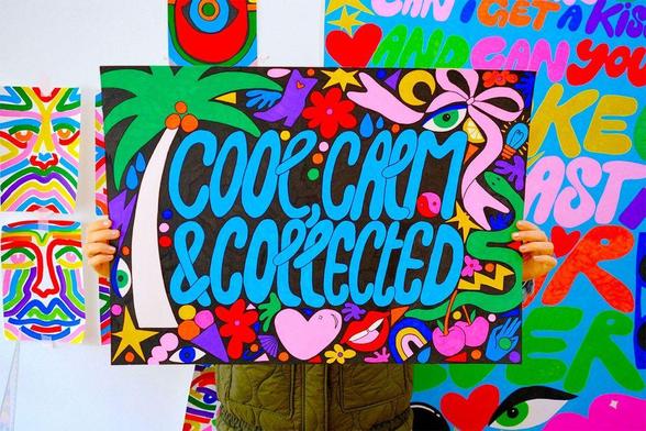 Person holding a colorful sign with the words "Cool, Calm & Collected" surrounded by abstract, vibrant illustrations and patterns. Other bold, graphic artworks are visible in the background.