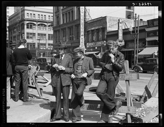 The image is a black-and-white photograph depicting several individuals gathered in an urban setting. The group consists of men, with one standing prominently on the left side wearing a dark suit and hat, while two others are conversing closely at center stage; both also dressed formally but without hats. One man leans against a wooden plank, smoking or contemplating something, evidenced by his hand near his face in a thoughtful gesture.

Behind them is a street filled with pedestrians and vehicles indicative of an earlier era, likely mid-20th century based on the fashion styles and automobile design visible. Various storefronts line the sidewalk including signs for businesses like "HONEY LOAN" indicating financial services available at that time, as well as establishments such as cafes.

The architecture suggests a downtown area with multi-story buildings featuring commercial signage in bold lettering above their entrances. Some of these are illuminated by electric lights which were not common earlier than late 19th century or early 20th-century America. The photograph exudes an atmosphere reflective of urban life during the Great Depression era, capturing both social interaction and economic struggle within a bustling cityscape.

This image was taken in San Francisco, California according to provided information linked via alt text which also references "Salvation Army" indicating possible charitable or community invol [...]