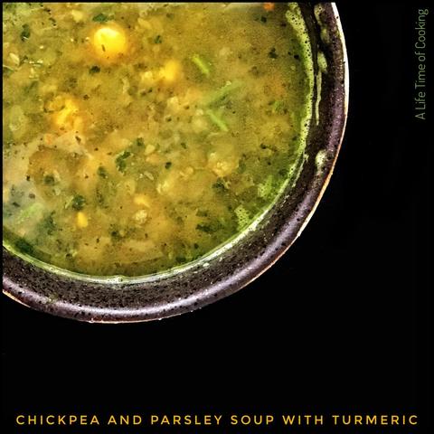 A close-up view of a bowl of soup, prominently displayed against a black background. The soup is in a dark, textured ceramic bowl, with a visible rim that has a rough, earthy appearance. The soup itself has a light greenish-yellow colour, indicating the presence of turmeric, and contains visible chunks of chickpeas and parsley, along with other ingredients that suggest a hearty, vegetable-based dish.