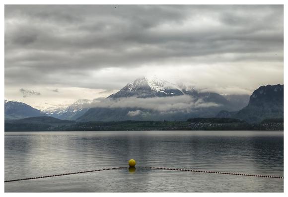 A still lake, you can see the patterns of the raindrops on the water. Mountains beyond the shore. Grey rain clouds.