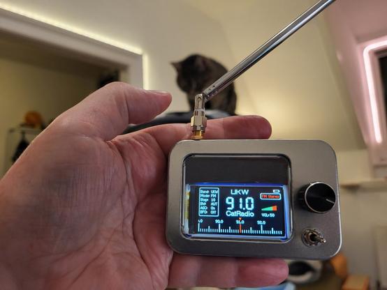 In der Hand ein kleines Radio in einer Blechdose. Empfängt 91 MHz Catradio. Im Hintergrund unscharf und dunkel Tigerkatze Lissy.