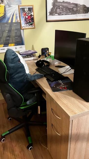 A playful setup featuring a large blahaj shark plush sitting in a gaming chair at a wooden desk. The desk is equipped with a computer monitor, keyboard, and gaming mouse pad. Surrounding decor includes a calendar and framed artwork, with gaming accessories visible on the desk.