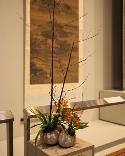 A minimalist ikebana arrangement in three stone-like vases featuring tall bare branches and small clusters of orchids and greenery, displayed in front of a hanging traditional East Asian scroll painting.