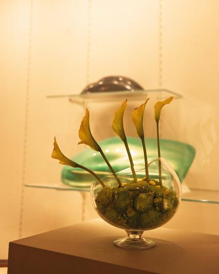 A delicate arrangement of yellow calla lilies and green moss-like filler in a round broken glass vessel, standing in front of abstract green glass art.