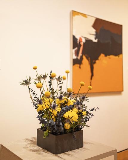 A vibrant floral arrangement of yellow and deep blue flowers in a black rectangular container, placed near an abstract orange and black painting.