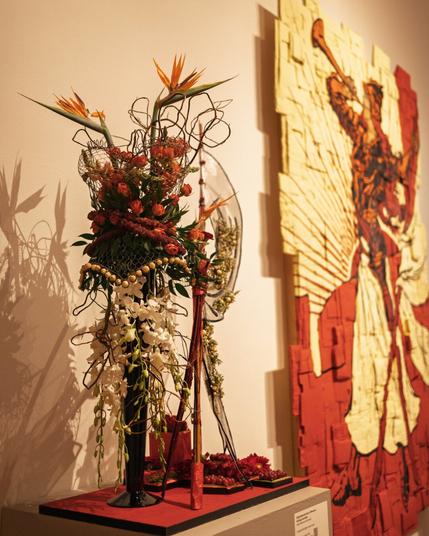 A dramatic and tall floral sculpture with red, orange, and white flowers, birds of paradise, and decorative wire elements, echoing a dynamic red-toned artwork of a figure with a trumpet.