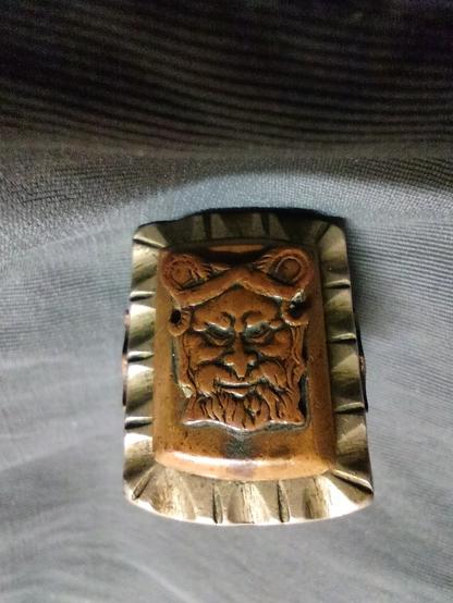 metal (maybe brass) ring with a carved bearded face on a brownish rectangular base.  the hair is parted and curls upward on either side, giving the appearance of horns.