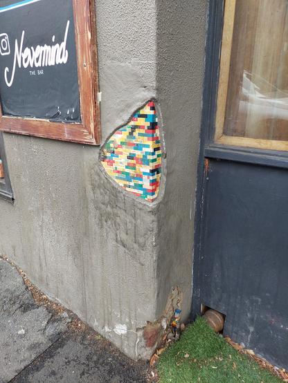 A wall rendered in grey concrete. A small section has no render on it revealing small pieces of random colour like a mosaic