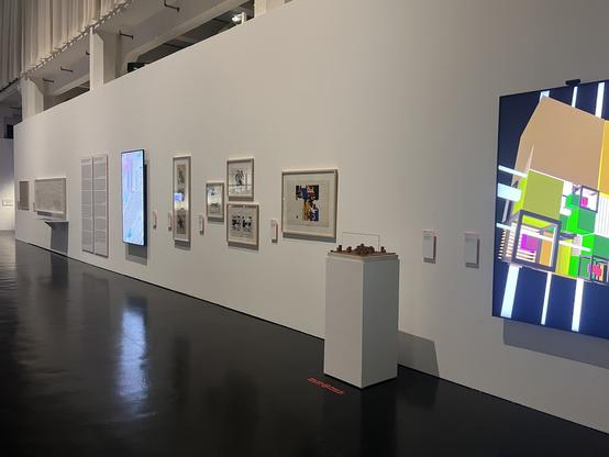 Photo of a large gallery space with printed and screen-based abstract artworks and info panels mounted on a long white wall. One of the pieces (titled "De/Frag", by the author) is shown on a large 75" screen mounted in portrait format, its light reflecting on the shiny black floor. The other screen on the right-hand side shows a 3D composition of cubes (artist unknown to me).  Image credit: Clara Runge
