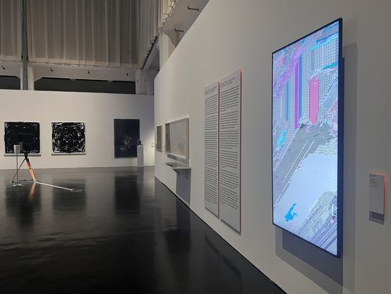 Photo of a large gallery space with artworks and info panels mounted on long white walls. One of the pieces in the foreground (titled "De/Frag", by the author) is shown on a large 75" screen mounted in portrait format, its light reflecting on the shiny black floor. Image credit: Clara Runge