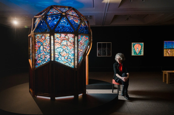 Photo from the linked NYTimes article. The original alt text in the article is: "The artist Judith Schaechter sits on a stool outside her colorful dome. Stained glass panels are displayed on a wall behind her."