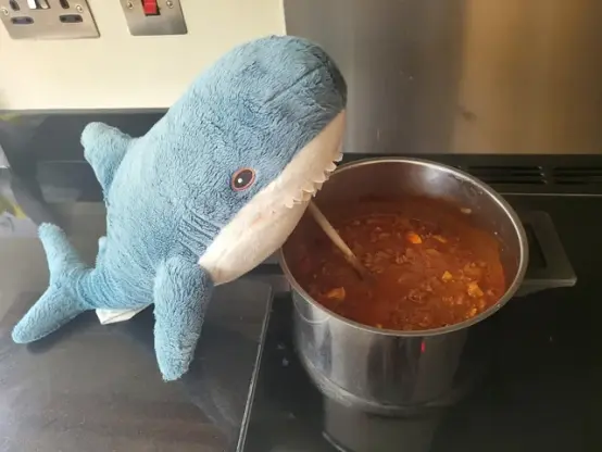 This image shows the IKEA stuffed animal blahaj preparing a soup dish in a big pot almost acting as a chef!