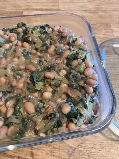 A glass container filled with a mixture of cooked white beans, chopped greens, and translucent vermicelli, all combined in a savory sauce. The container is placed on a wooden surface.