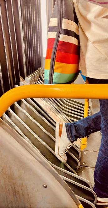 A colorful scene in the interior of an articulated bus in NYC. At right frame are the lower torso and legs of a person facing right, wearing a light-colored jacket, blue jeans, and white sneakers with brown trim. The left leg is raised with the shoe resting on the accordion bellows between the two sections of the bus. The person carries a large zippered shoulder bag with wide stripes of many colors, from black and white at the top, to reds and oranges in the middle, to blues and greens on the bottom. In the foreground is a yellow horizontal guardrail that bisects the image and curves slightly downward at left frame.