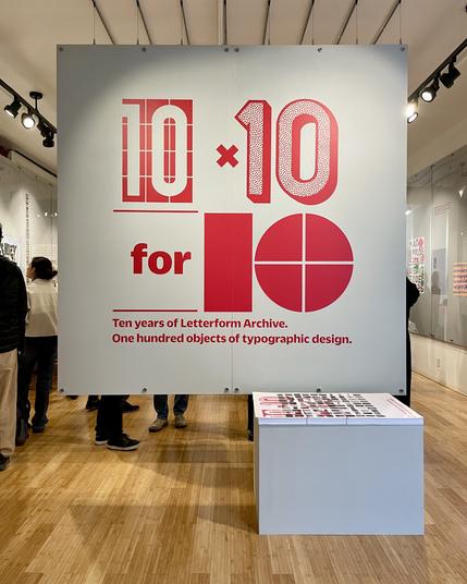 Photo of a wood-floored gallery with a large floating gray wall filling most of the frame. In the wall are large red vinyl letters: “10 × 10 for 10” in different styles, and “Ten years of Letterform Archive. One hundred objects of typographic design.” in smaller text. A stack of posters on a pedestal is in the foreground, and people looking at art in the gallery in the background.