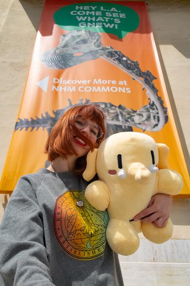 Photo of Lety and the Creature Mastodon stuffed toy beneath a giant promotional banner outside the Natural History Museum of Los Angeles County on a bright, sunny day. From top to bottom, Lety is seen wearing a gray graphic sweater featuring a rainbow seal of the Kennedy Space Center. She's standing in the foreground, holding the Creature and smiling down at the camera. Above and behind them both is a giant promotional banner featuring an image of Gnatalie the Green Dino for the new wing at the museum.

A basic transcript of the banner follows.

HEY L.A. COME SEE WHAT'S GNEW!

Discover More at NHM COMMONS