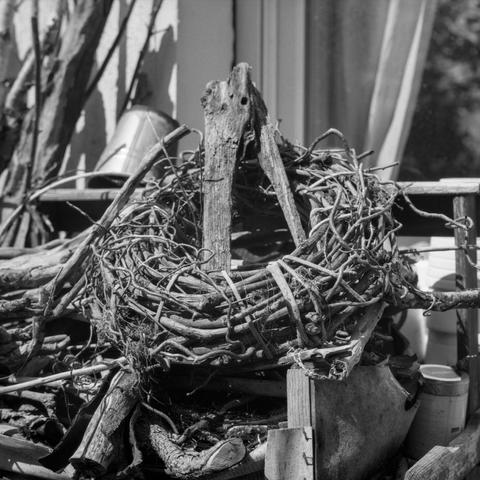 Black-and-white photo of a rustic wreath made of twisted vines and branches, resting amid a pile of old wood and garden tools.