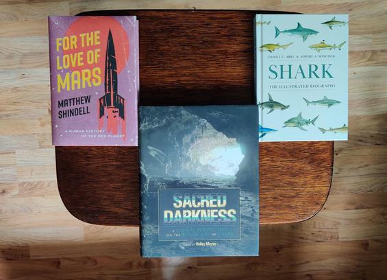 A top-down photo of three books lying down on a small, brown, wooden table. On the left, For the Love of Mars, showing a cartoony drawing of a black rocket lifting off. In the middle, Sacred Darkness, showing the brightly lit opening of a cave mouth, photographed from inside a large cave with a wide and flat floor. On the right, Shark: The Illustrated Biography, showing some 10 colour illustrations of different shark species. The background shows a beech-coloured laminate floor.