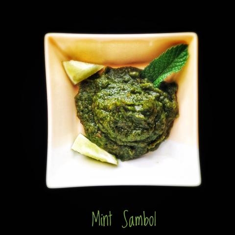 A green chutney in a square bowl, with a mint leaf and wedges of lime.