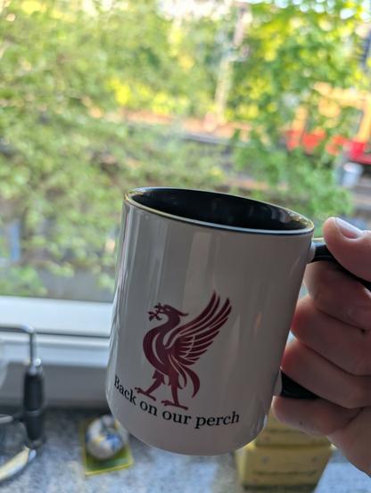 A hand Holding a white coffee mug in Front oft a window. The mug got a red liverbird and the text: back on our perch