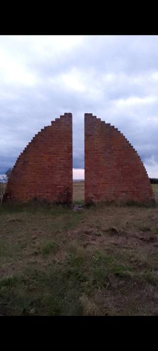 Two red-brown brick pilars that together form half of a circle, with some space between them where you can see the ocean behind them and the sky is cloudy.