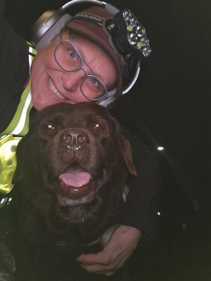 Night time selfie of a person and a chocolate brown Labrador with greying snout. The person is wearing a black jacket, yellow safety harness, red cap, silver headphones and blue-rimmed glasses. On their cap rests a headstrap torch in off position. Their free hand is around the dog's shoulders. Both are facing the camera. The dog's mouth is slightly open, showing off some blep. The person is smiling, their head resting on the dog's head. She's grateful for the time spent together.