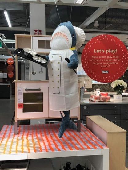 At IKEA, a BLÅHAJ shark plushie dressed as a chef complete with white uniform and chef's hat, stands proudly holding a spatula next to a pink toy kitchen set. A bright red circular sign reads "Let's play! Make lunch, play shop or create a puppet show? Let your imagination decide!" The shark appears to be demonstrating how children can use IKEA's play furniture for imaginative play. The display is set on a colorful orange and white striped platform, with additional kitchen play accessories visible nearby. BLÅHAJ has traded swimming for cooking in this charming store display that encourages creative play.​​​​​​​​​​​​​​​​