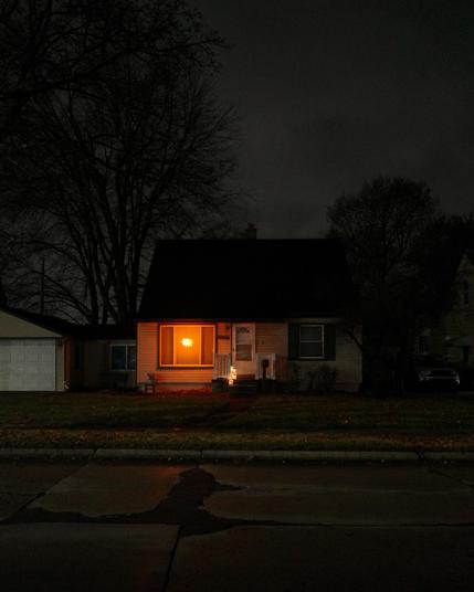 Color photograph looking at the front of a small bungalow house on an overcast night. There's a brightly shining decorative light hanging in the picture window to the left of the front door giving off an orange light while everything around is in darkness and shadow, trees silhouetted against the gray overcast sky