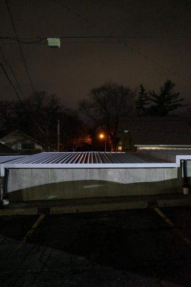 Color photograph in a parking lot at night, looking across an empty parking spot on an incline and over a concrete embankment that forms part of the level top of a carport to a parking lot on lower ground, beyond which are darkened apartment buildings and the tops of some trees silhouetted against an overcast sky glowing orange ish gray from light pollution, a lone streetlight shining in the darkness against trees enclosing it like a cave